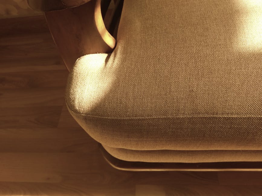Close-up image of a beige upholstered armchair with textured fabric, showing the seat cushion and rounded armrest. The chair is situated on a wooden floor with a natural finish, and part of a wooden side table is visible at the top left corner. The fabric appears clean and well-maintained, with even stitching and no visible stains or dust. Soft, warm lighting highlights the smooth surface and subtle texture of the upholstery. The setting suggests a tidy, comfortable living space, consistent with professional domestic cleaning standards. The overall scene reflects attention to surface cleaning and upholstery care by Carpet Cleaning Swiss Cottage as part of their deep cleaning and sanitisation services showcased in the Finchley Road upholstery cleaning before and after case study.