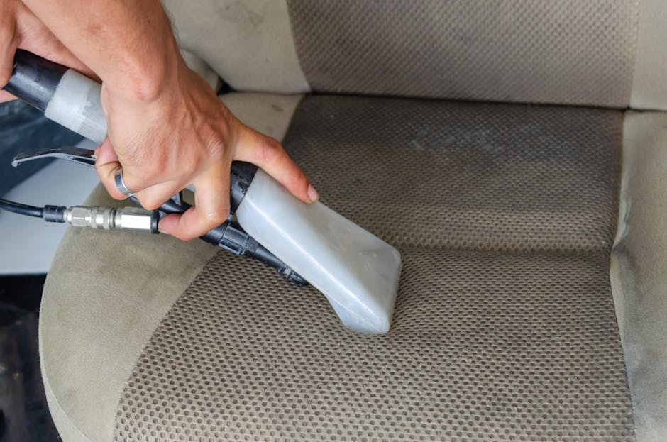 A person holding a black vacuum cleaner with a flexible hose is performing deep cleaning or surface sanitisation on a yellow and white patterned cushion placed on a dark fabric sofa in a modern living room. The sofa is positioned against a plain light-colored wall, with bright, even lighting highlighting the cleanliness and well-maintained condition of the furniture. The scene showcases domestic upholstery cleaning processes, indicating professional efforts by Carpet Cleaning Swiss Cottage to enhance hygiene and cleanliness in the home setting, as referenced in the Finchley Road upholstery cleaning before and after case study.