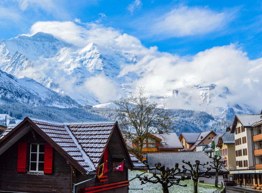 A picturesque Swiss village during winter, featuring traditional wooden houses with dark brown facades, red shutters, and snow-covered tiled roofs. Behind the houses, a backdrop of majestic snow-capped mountains rises into a partly cloudy sky, with patches of blue peeking through. Bare trees with twisted branches line the streets, which are lined with modern multi-storey buildings. The bright winter sunlight illuminates the scene, highlighting the pristine snow on rooftops and the vibrant colors of the houses, creating a serene, clean, and inviting atmosphere typical of a mountain resort village. This image exemplifies the importance of surface cleaning and meticulous maintenance to preserve the aesthetic appeal of properties in snowy environments, aligning with the themes of sanitisation and domestic cleaning promoted by Carpet Cleaning Swiss Cottage.