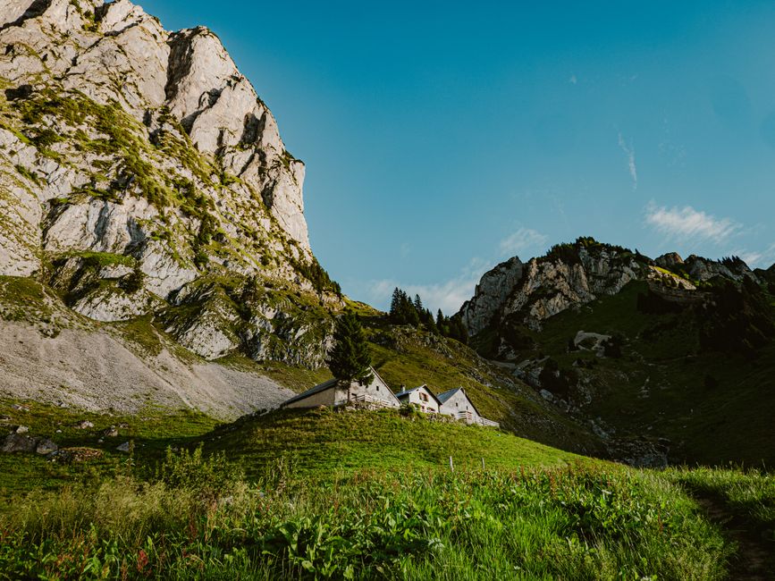 A rural landscape featuring several white houses with dark roofs situated at the base of towering, rugged limestone mountains with patches of green vegetation, including trees and grass, under a clear blue sky with a few wispy clouds, illuminated by bright sunlight, illustrating a peaceful countryside scene associated with domestic and outdoor cleaning environments. This image reflects the natural setting often encountered during outdoor maintenance and surface cleaning activities, aligning with the themes of hygiene and environmental care promoted by Carpet Cleaning Swiss Cottage, as seen on their website offering same day quotes and prices for cleaning services.