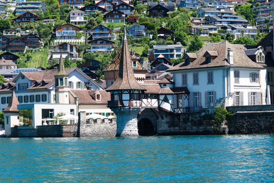 A scenic view of a lakeside residential area with a historic chalet-style building featuring a spire and dark wood accents. Surrounding the building are white and pastel-colored houses with tiled roofs, some with decorative shutters and large windows. These structures are situated along the water's edge, with a stone wall and a small arched tunnel under the main building. In the background, numerous modern homes with large glass balconies and sloped roofs are visible on the hillside, set amidst lush green vegetation. The scene is well-lit, emphasizing the clean and well-maintained appearance of the buildings, with calm blue water in the foreground reflecting the structures and sky.